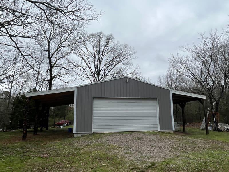 Gray steel building with white trim and open carport wings, built by Speedy Gonzalez in a wooded Garland County lot